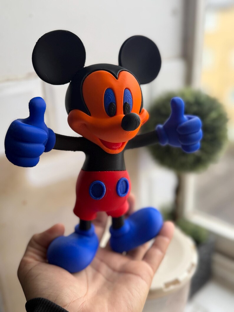Pu&ograve; includere: Una colorata statuina di Topolino, con orecchie nere, corpo nero, viso arancione, occhi blu, pantaloncini rossi e guanti e scarpe blu. La figura fa il gesto del pollice in su con entrambe le mani. La statuina &egrave; tenuta in una mano.