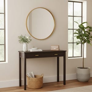 Puede incluir: Una consola marrón oscuro con dos cajones y herrajes dorados. Un espejo redondo con marco dorado cuelga sobre la mesa. Una cesta de mimbre, un jarrón con flores y un pequeño bolso están sobre o cerca de la mesa.
