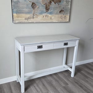 May include: A white wooden console table with two drawers and black hardware. Above the table hangs a framed world map with a distressed, textured design in shades of brown, blue, and beige.