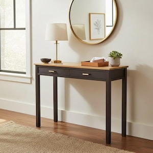 May include: A modern console table with two drawers, a light wood top, and dark brown legs. A gold-toned lamp, a small bowl, and a decorative tray with a plant sit on top. A round gold mirror hangs on the wall.