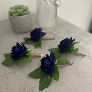 May include: Four navy blue rose boutonnieres with green leaves and twine-wrapped stems are arranged on a grey surface. A small succulent in a grey pot and a decorative glass bottle are in the background.