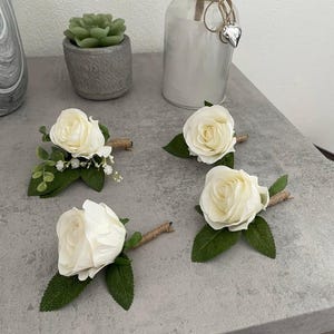 May include: Four white rose boutonnieres with green leaves and twine stems are arranged on a grey surface. The roses are the focal point, with detailed petals. The boutonnieres are likely for a wedding or special event.