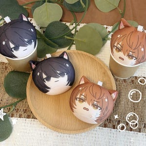 May include: Four plush cat-faced keyrings with anime-style features. Two are black and two are brown, each with a different expression. They are arranged on a wooden plate and in paper cups, surrounded by greenery and decorative elements.