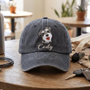 Puede incluir: Una gorra de béisbol de algodón lavado gris carbón con un bordado de un perro Schnauzer con la lengua roja y el nombre Cody cosido en letra cursiva blanca debajo de la ilustración del perro en el panel frontal de la gorra.