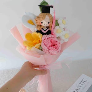 May include: A crocheted bouquet featuring a graduation doll with a black cap and pink tassel, surrounded by crocheted flowers in white, yellow, and pink. The bouquet is wrapped in pink paper and tied with a pink ribbon.