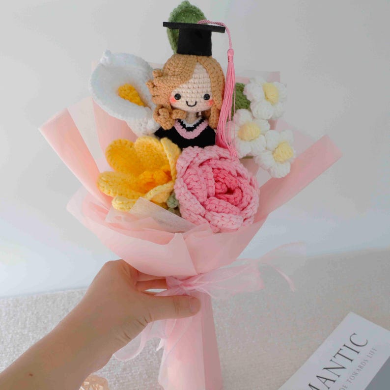 May include: A crocheted bouquet featuring a graduation doll with a black cap and pink tassel, surrounded by crocheted flowers in white, yellow, and pink. The bouquet is wrapped in pink paper and tied with a pink ribbon.