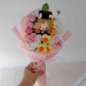 May include: A handmade crochet bouquet featuring a graduate doll with a black cap and yellow tassel, surrounded by pink, white, and yellow crocheted flowers. The bouquet is wrapped in pink paper and tied with a sheer ribbon.