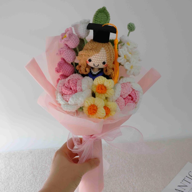 May include: A handmade crochet bouquet featuring a graduate doll with a black cap and yellow tassel, surrounded by pink, white, and yellow crocheted flowers. The bouquet is wrapped in pink paper and tied with a sheer ribbon.