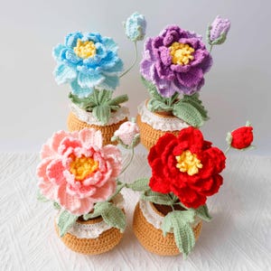 May include: Four crocheted flowers in individual tan pots, each featuring a white lace trim. The flowers are blue, purple, pink, and red, all with yellow centers and green leaves. Each pot includes a small, unopened flower bud on a thin stem.