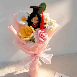 May include: A crocheted bouquet featuring a graduation doll wearing a black cap and gown, surrounded by crocheted flowers in white, yellow, and pink. The bouquet is wrapped in pink paper and tied with a sheer ribbon.
