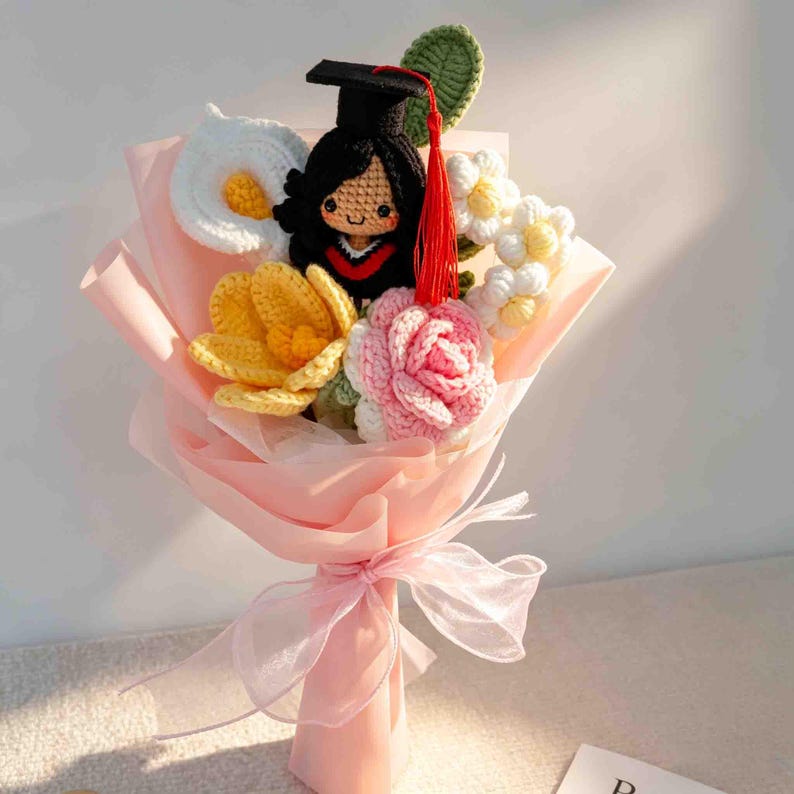 May include: A crocheted bouquet featuring a graduation doll wearing a black cap and gown, surrounded by crocheted flowers in white, yellow, and pink. The bouquet is wrapped in pink paper and tied with a sheer ribbon.