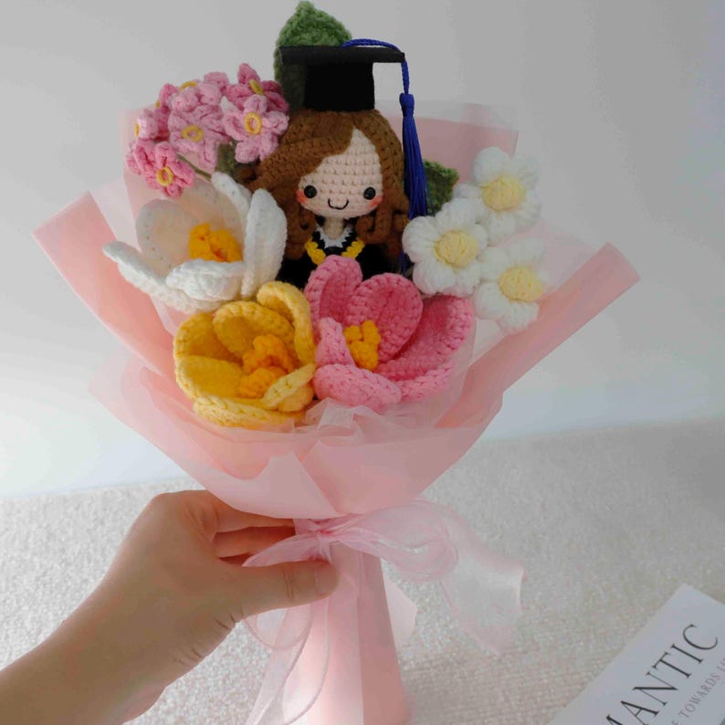 May include: A handmade crochet bouquet featuring a graduation doll wearing a black cap and gown, surrounded by colorful crochet flowers in pink, white, and yellow. The bouquet is wrapped in pink paper and tied with a ribbon.