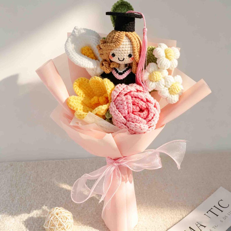 May include: A crocheted bouquet featuring a graduation doll wearing a black cap and gown, surrounded by crocheted flowers in white, yellow, and pink. The bouquet is wrapped in pink paper and tied with a matching ribbon.