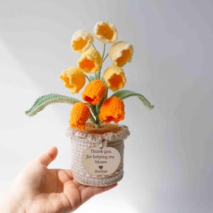 May include: A crocheted potted plant featuring yellow and orange bell-shaped flowers with green leaves. The beige pot has a scalloped rim and a wooden tag attached that reads: Thank you for helping me bloom, with a heart icon and the name Amber.