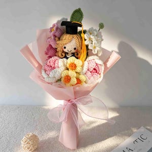May include: A handmade crochet bouquet featuring a doll wearing a graduation cap. The bouquet includes pink, white, and yellow crocheted flowers, wrapped in pink paper and tied with a pink ribbon. A unique graduation gift.