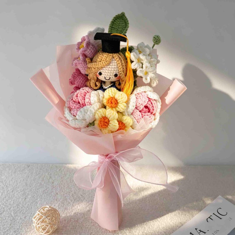 May include: A handmade crochet bouquet featuring a doll wearing a graduation cap. The bouquet includes pink, white, and yellow crocheted flowers, wrapped in pink paper and tied with a pink ribbon. A unique graduation gift.