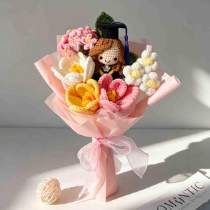 May include: A crocheted bouquet featuring a graduation doll with a black cap and gown, surrounded by colorful crocheted flowers in shades of pink, yellow, and white. The bouquet is wrapped in pink paper and tied with a sheer ribbon.
