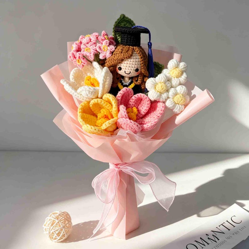May include: A crocheted bouquet featuring a graduation doll with a black cap and gown, surrounded by colorful crocheted flowers in shades of pink, yellow, and white. The bouquet is wrapped in pink paper and tied with a sheer ribbon.