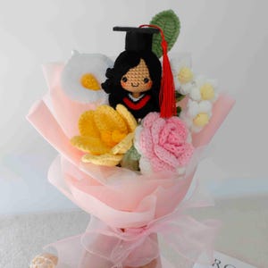 May include: A handmade crochet bouquet featuring a graduate doll wearing a black cap and gown, surrounded by crochet flowers in yellow, white, and pink. The bouquet is wrapped in pink paper and tied with a pink ribbon.