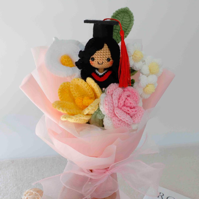 May include: A handmade crochet bouquet featuring a graduate doll wearing a black cap and gown, surrounded by crochet flowers in yellow, white, and pink. The bouquet is wrapped in pink paper and tied with a pink ribbon.