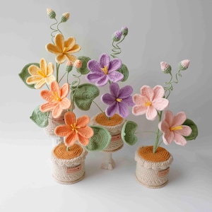 May include: Three potted crochet flower arrangements featuring yellow, orange, purple, and pink blossoms with green leaves and stems. Each plant is housed in a textured beige crocheted pot with a brown top and a small rectangular label on the side.
