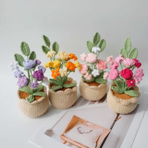 May include: Four crocheted potted plants featuring colorful flowers in shades of purple, yellow, orange, and pink. Each plant has green crocheted leaves and is housed in a beige, textured, round crocheted pot with a scalloped top edge.