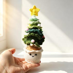 May include: A crocheted Christmas tree with tiered green branches, small colorful pom-pom ornaments, and a yellow star topper. The tree is planted in a cream-colored crocheted pot featuring a simple embroidered face with black eyes and a smile.