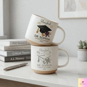May include: Two stacked white ceramic mugs with a speckled finish and tan base. Each mug features a graduation cap graphic, the text "Phinished PhD", and the name "Dr. Sophia, University of California 2026".