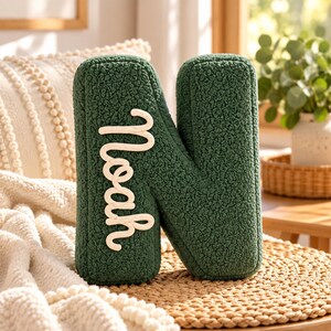 May include: A dark green letter N shaped cushion made of textured sherpa fabric. The letter features the name Noah written in a white, raised, cursive rope-style font vertically along the left side of the letter. The cushion has a soft, plush appearance.