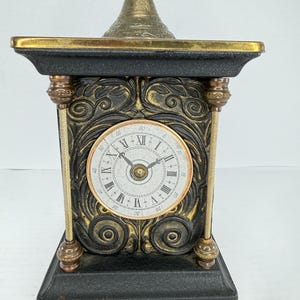Puede incluir: Un reloj antiguo negro y dorado con una esfera blanca con números romanos. El reloj tiene detalles ornamentados, detalles dorados y una parte superior decorativa. El reloj mide aproximadamente 30 cm de alto.