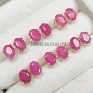 Puede incluir: Una colección de pendientes de botón con piedras preciosas rosas de forma ovalada montadas en garras de plata. Cada pendiente presenta una superficie facetada y está dispuesto en pares. El texto JEWELSBYLALITA es visible en la caja blanca.