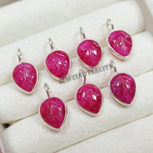 Könnte beinhalten: Sieben kleine, tropfenförmige Anhänger mit geschnitzten roten Edelsteinblättern in silberfarbenen Metallfassungen. Jeder Anhänger hat oben einen kleinen silbernen Biegering zur Befestigung. Der Text JEWELRYLALITA ist in der Mitte sichtbar.
