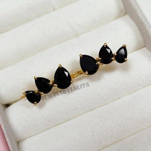 May include: A gold ear climber earring featuring five pear-cut black stones arranged in a graduated row. The metal setting holds each stone with small prongs. The text JEWELSBYLALITA is printed in grey across the center of the earring design.