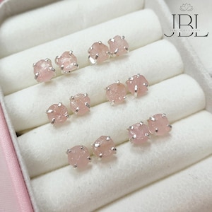 Può includere: Una collezione di orecchini a lobo in quarzo rosa con montatura color argento. Gli orecchini sono di colore rosa chiaro e hanno una consistenza ruvida e naturale. Gli orecchini sono esposti in un portagioie bianco.