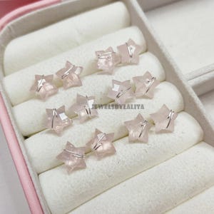 Op de afbeelding: Roze ster-vormige oorbellen, elk vastgezet met zilverdraad, worden gepresenteerd in een roze en witte sieradendoos. De tekst "JEWELSBYLALITA" is zichtbaar.