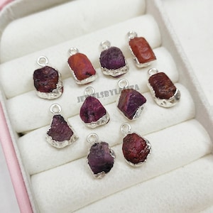 Peut inclure: Un ensemble de neuf pendentifs en rubis brut rouge foncé, chacun serti dans une monture en métal argenté avec une petite boucle circulaire sur le dessus pour la fixation. Les pierres varient en forme et en taille, avec des textures naturelles.