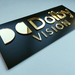 May include: A rectangular black sign featuring the gold-colored, reflective, three-dimensional text "Dolby Vision" mounted on the surface. The letters are raised, creating a layered effect against the smooth, matte black background panel.
