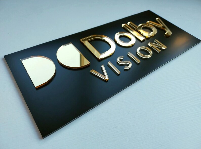 May include: A rectangular black sign featuring the gold-colored, reflective, three-dimensional text "Dolby Vision" mounted on the surface. The letters are raised, creating a layered effect against the smooth, matte black background panel.
