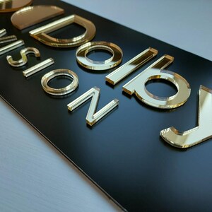 May include: A rectangular black sign featuring the raised, reflective gold-toned acrylic text "DOLBY VISION" in a sans-serif font. The letters are mounted on a smooth, matte black surface, creating a high-contrast, three-dimensional appearance.