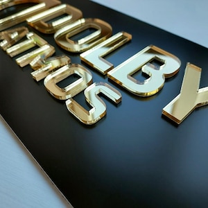 May include: A rectangular black sign featuring the raised, reflective gold-colored acrylic text DOLBY ATMOS. The letters are cut in a bold, sans-serif font and are mounted onto the smooth, matte black surface, creating a high-contrast appearance.