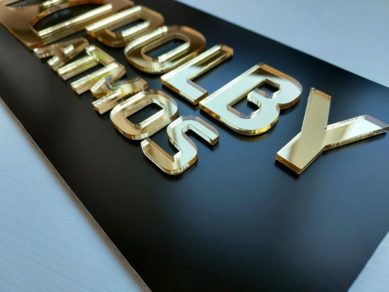 May include: A rectangular black sign featuring the raised, reflective gold-colored acrylic text DOLBY ATMOS. The letters are cut in a bold, sans-serif font and are mounted onto the smooth, matte black surface, creating a high-contrast appearance.