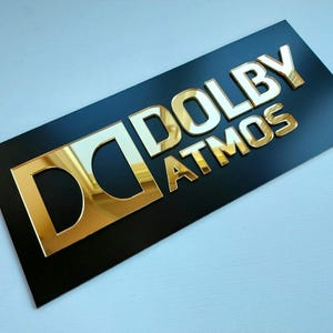 May include: A rectangular black sign featuring the gold-colored, reflective Dolby Atmos logo and text. The design includes the iconic double-D symbol followed by the word DOLBY and the word ATMOS positioned directly underneath in capital letters.