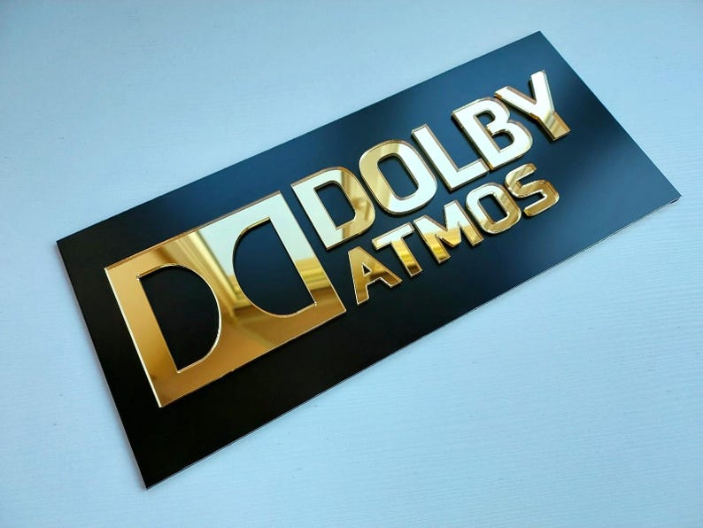 May include: A rectangular black sign featuring the gold-colored, reflective Dolby Atmos logo and text. The design includes the iconic double-D symbol followed by the word DOLBY and the word ATMOS positioned directly underneath in capital letters.