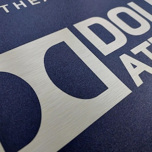 May include: Close-up of a navy blue surface with the word "DOLBY ATMOS" in large, white, sans-serif letters. The "D" and the "O" of "DOLBY" are partially obscured by a brushed silver-colored rectangle. The word "THE" is visible in white at the top.