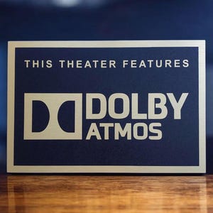 May include: A rectangular sign with a gold border and a navy blue center. The sign reads "THIS THEATER FEATURES" at the top, with the Dolby Atmos logo in gold below. The sign is on a wooden surface.