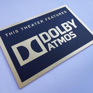 May include: A rectangular sign with a gold border and a navy blue center. The text "THIS THEATER FEATURES" is at the top, with "DOLBY ATMOS" in large, bold, gold letters below. The Dolby logo is also gold.