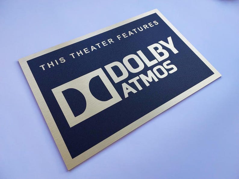 May include: A rectangular sign with a gold border and a navy blue center. The text "THIS THEATER FEATURES" is at the top, with "DOLBY ATMOS" in large, bold, gold letters below. The Dolby logo is also gold.
