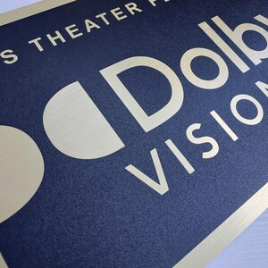 Może przedstawiać: Prostokątny znak z granatowym tłem i złotą obw&oacute;dką. Znak zawiera słowa "THIS THEATER PRESENTS" na g&oacute;rze i "Dolby VISION" dużymi, stylizowanymi literami. Widoczne jest r&oacute;wnież logo Dolby.