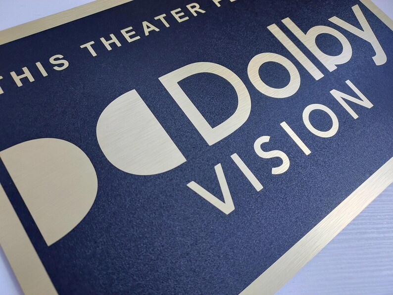 Może przedstawiać: Prostokątny znak z granatowym tłem i złotą obw&oacute;dką. Znak zawiera słowa "THIS THEATER PRESENTS" na g&oacute;rze i "Dolby VISION" dużymi, stylizowanymi literami. Widoczne jest r&oacute;wnież logo Dolby.