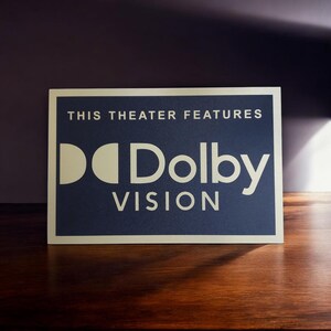Może przedstawiać: Prostokątny znak z napisem "THIS THEATER FEATURES" nad logo "Dolby VISION". Znak ma ciemnoniebieskie tło z białym tekstem i logo. Znak znajduje się na drewnianej powierzchni.